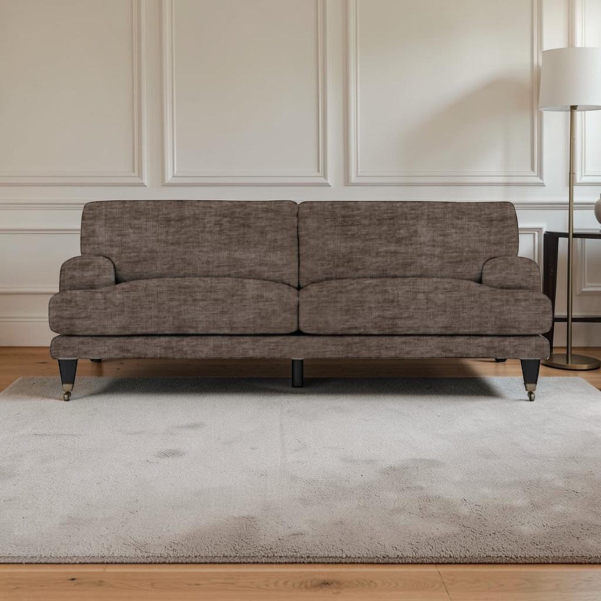 A upholstered sofa in textured taupe-brown fabric, shown in a styled living room setting with a light gray rug, wooden floor, white paneled wall, and a floor lamp.