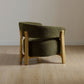 A modern upholstered accent chair with a curved olive green fabric seat and backrest, supported by light natural wood legs, shown on a light wood plank floor against a neutral beige wall.