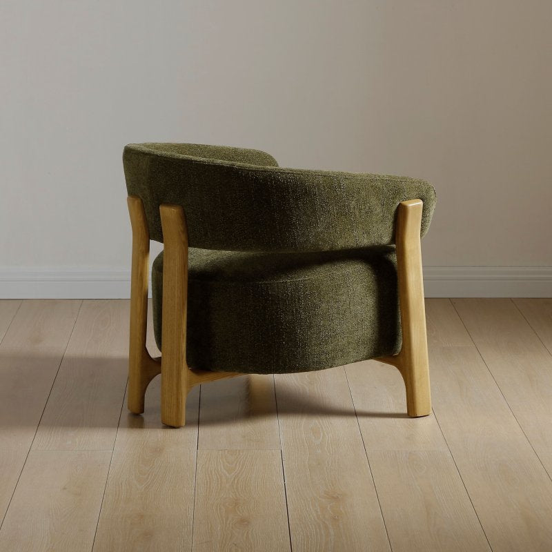 A modern upholstered accent chair with a curved olive green fabric seat and backrest, supported by light natural wood legs, shown on a light wood plank floor against a neutral beige wall.