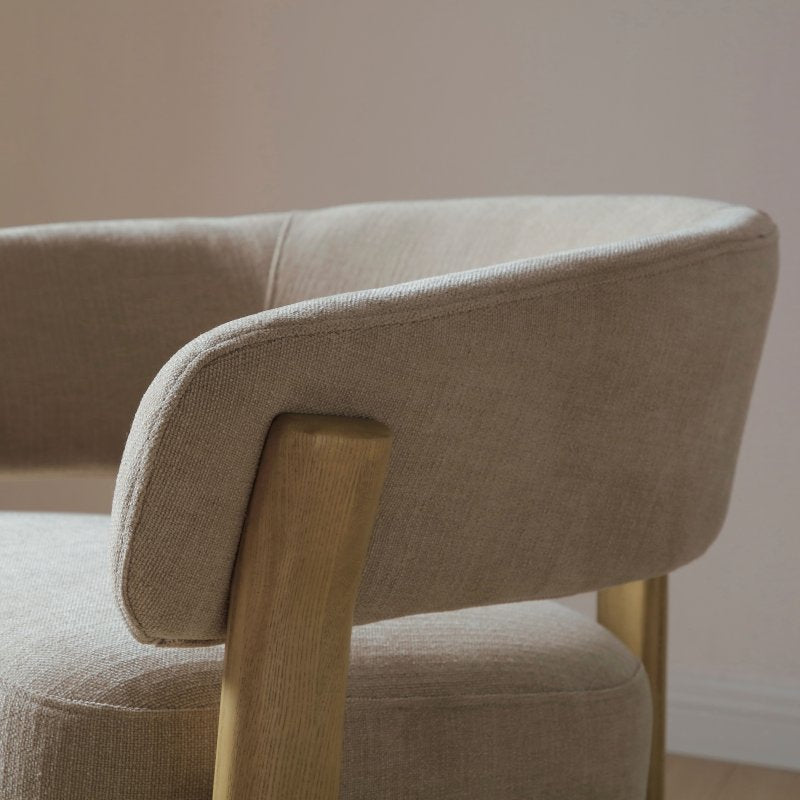 Beige upholstered chair with wooden armrest and legs against a neutral wall.