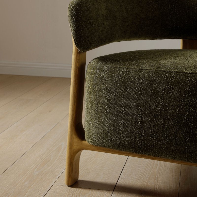 Close-up of a modern upholstered armchair with textured olive green fabric and a light natural wood frame, shown on a light wood floor against a neutral wall.