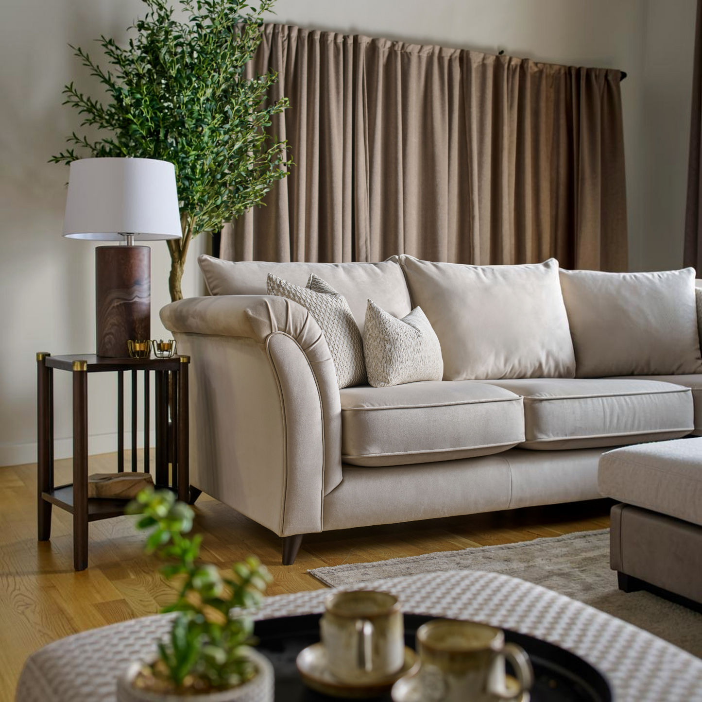 A cream-colored fabric sofa in a styled living room setting, featuring neutral beige curtains, a dark wood side table with a lamp, and a potted green tree.