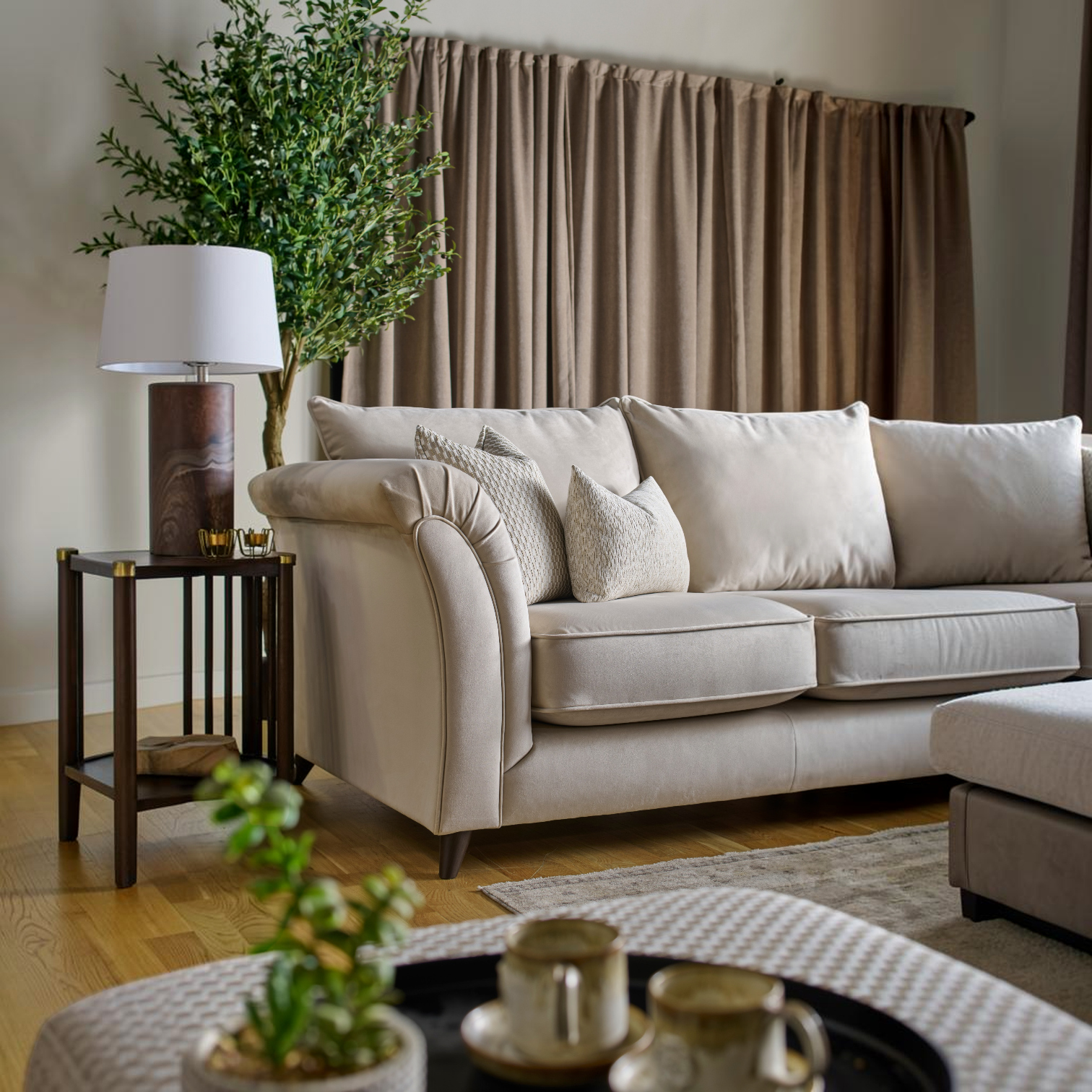 A cream-colored fabric sofa in a styled living room setting, featuring neutral beige curtains, a dark wood side table with a lamp, and a potted green tree.
