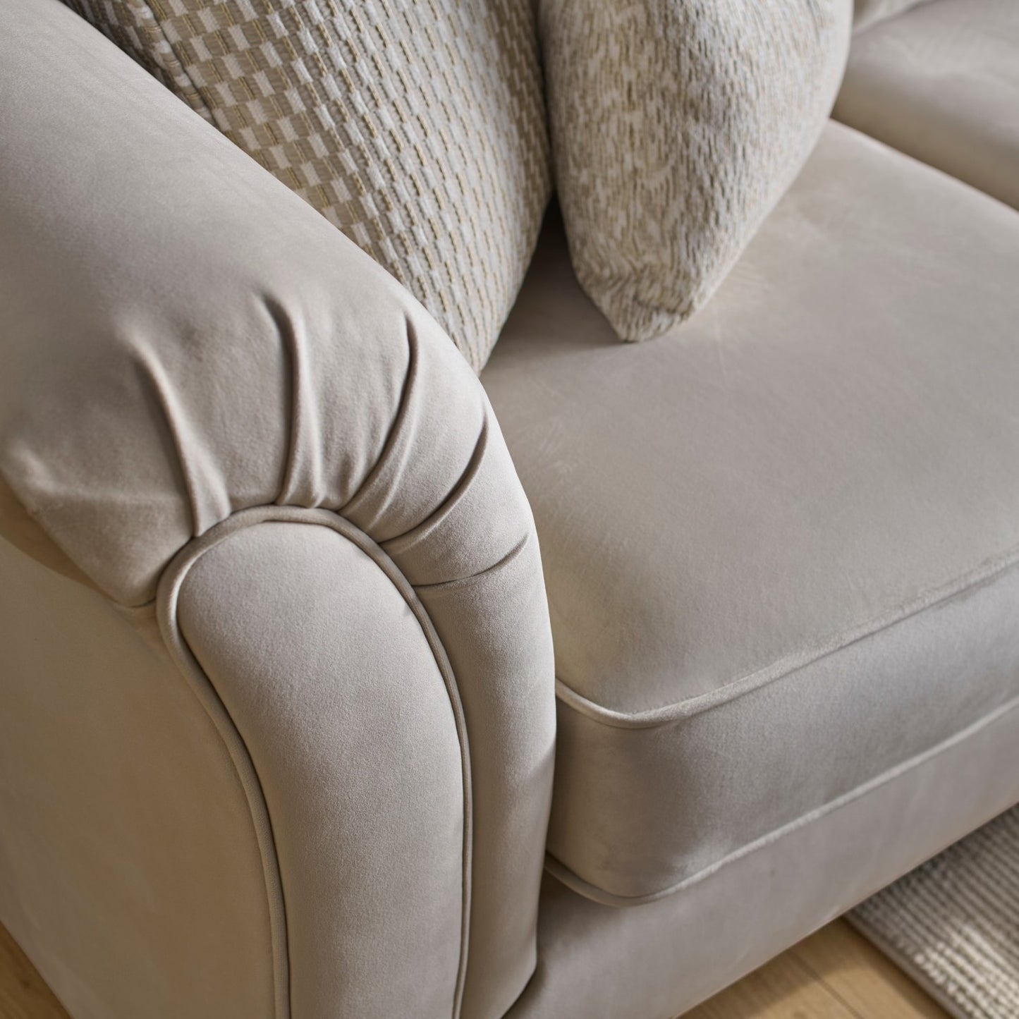 Close-up of a light beige upholstered sofa arm and seat cushion with two textured throw pillows, photographed indoors on a wood floor.