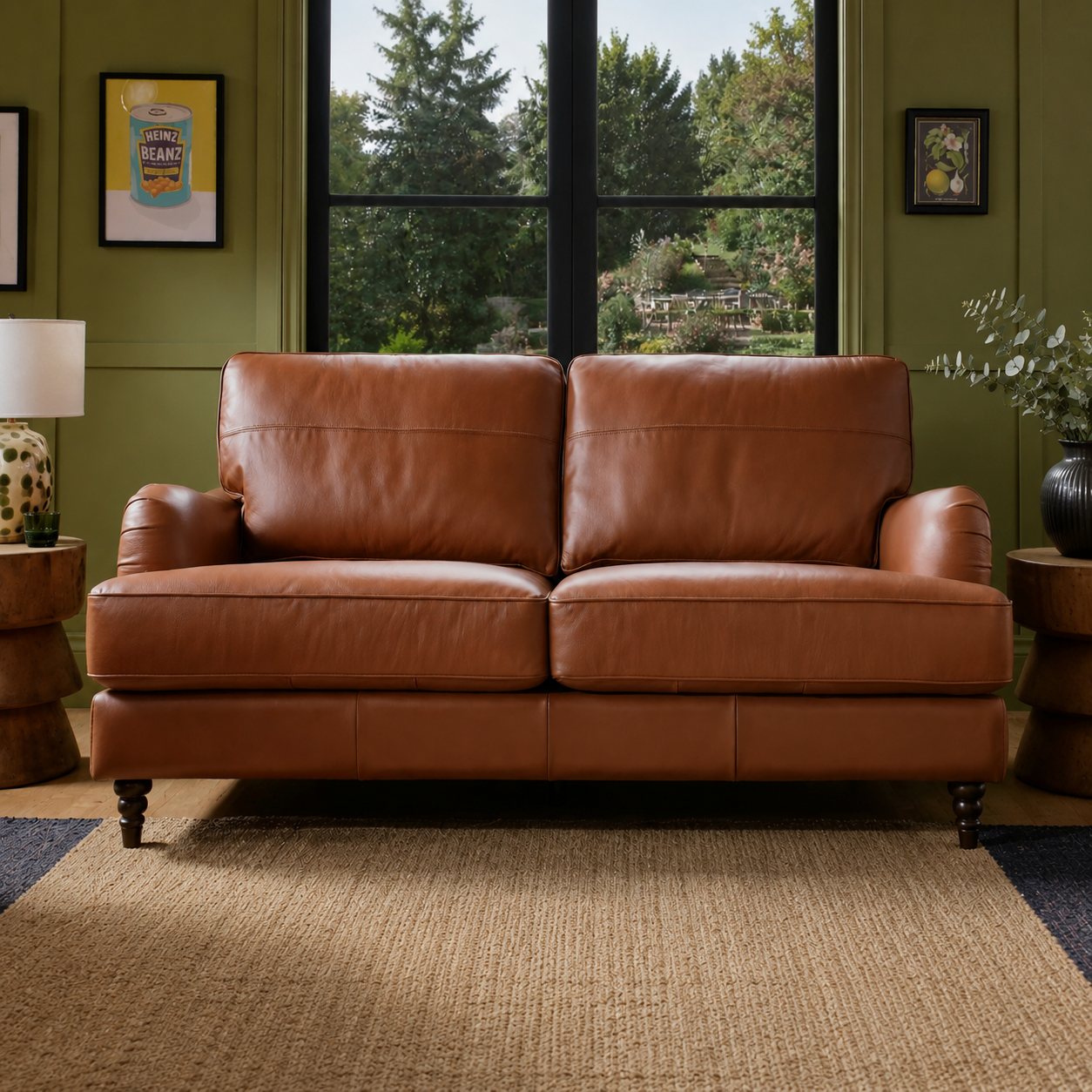 A two-seat brown leather loveseat sofa placed in a styled living room with olive green walls, a large window showing trees outside, wooden side tables, framed wall art, and a woven rug.