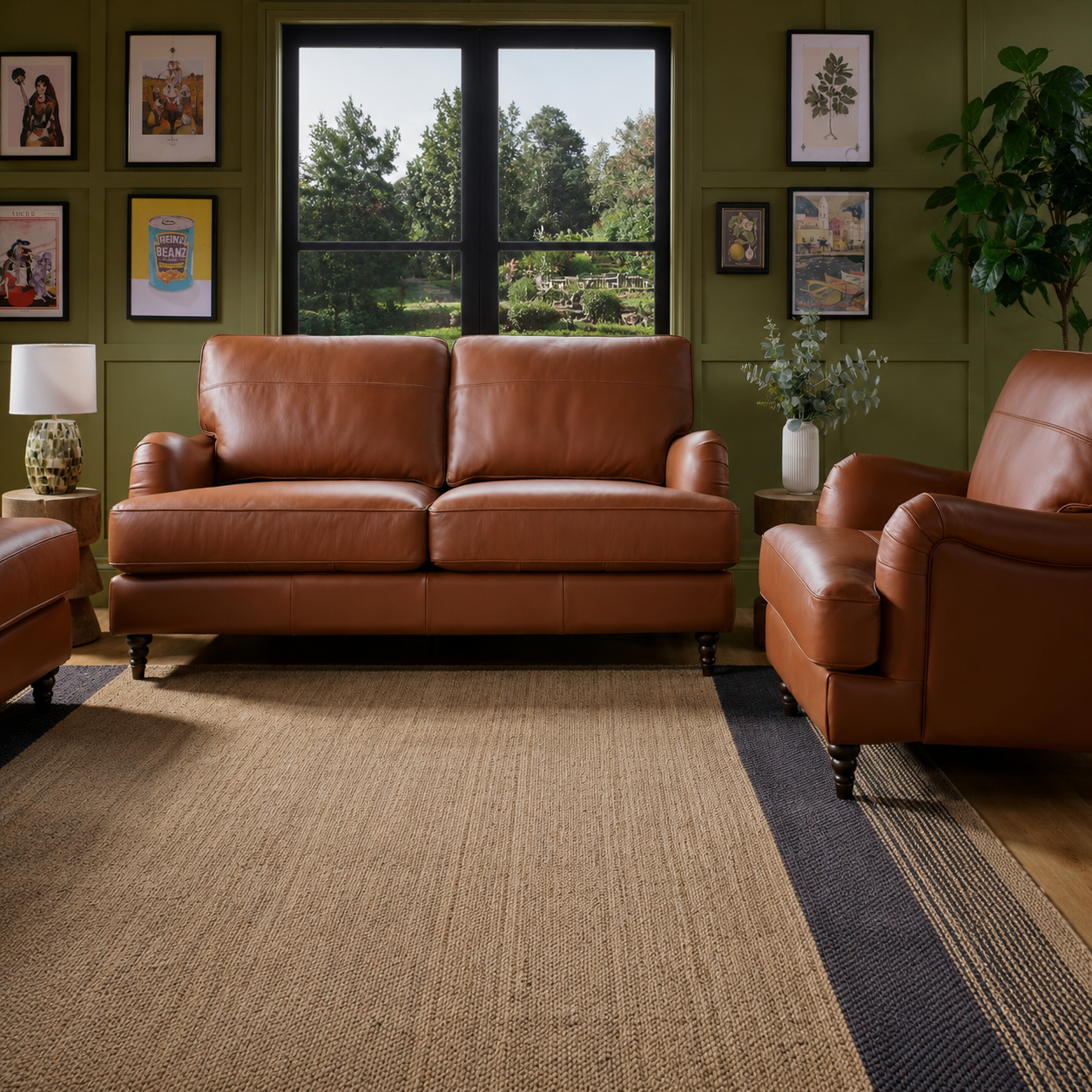 A brown leather sofa and matching armchair in a styled living room with olive green paneled walls, framed botanical art, a window view of trees, and a large woven area rug.