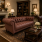 A classic tufted Chesterfield sofa upholstered in saddle tan leather, positioned in a richly decorated traditional living room with a dark wood coffee table, rug, bookshelves, and lamps.