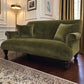 An olive green velvet sofa with two matching pillows, set in a classic room with dark wood floors, a patterned rug, a floor lamp, and gold curtains.