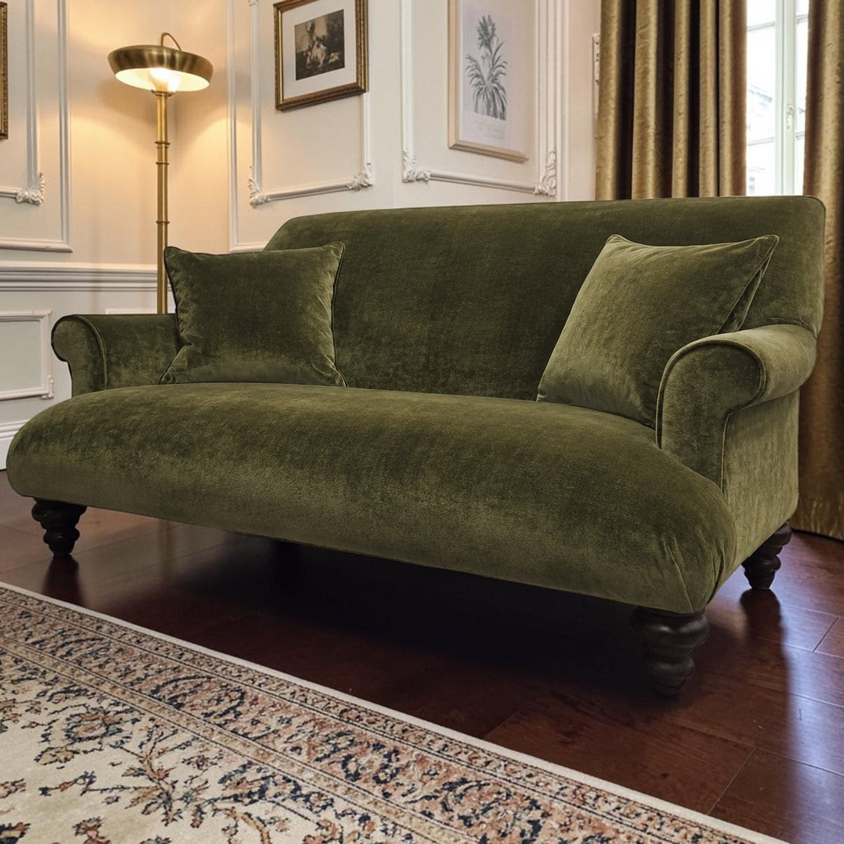 An olive green velvet sofa with two matching pillows, set in a classic room with dark wood floors, a patterned rug, a floor lamp, and gold curtains.