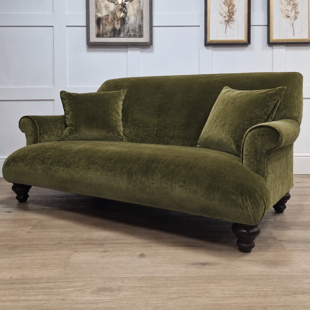 An olive green velvet sofa with dark wooden legs, styled in a showroom setting with a white paneled wall and light wood flooring.
