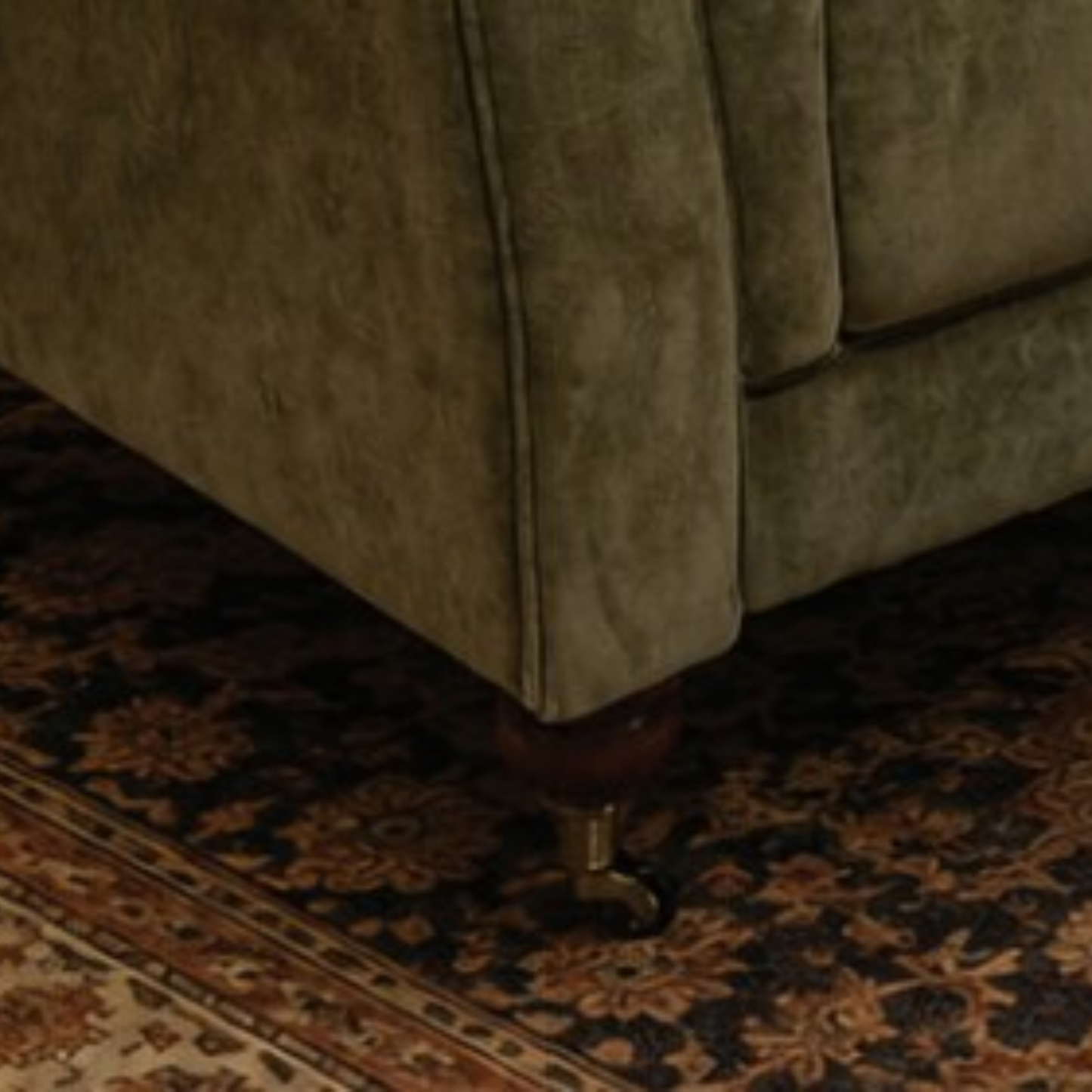 Close-up of the corner of an upholstered armchair or sofa in a muted olive green fabric, showing a dark wood leg with a brass castor wheel over a patterned area rug.