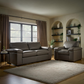 A staged living room featuring a leather sofa and a matching armchair placed on a large beige shag rug, with brown curtains, wood flooring, and built-in arched shelves in the background.