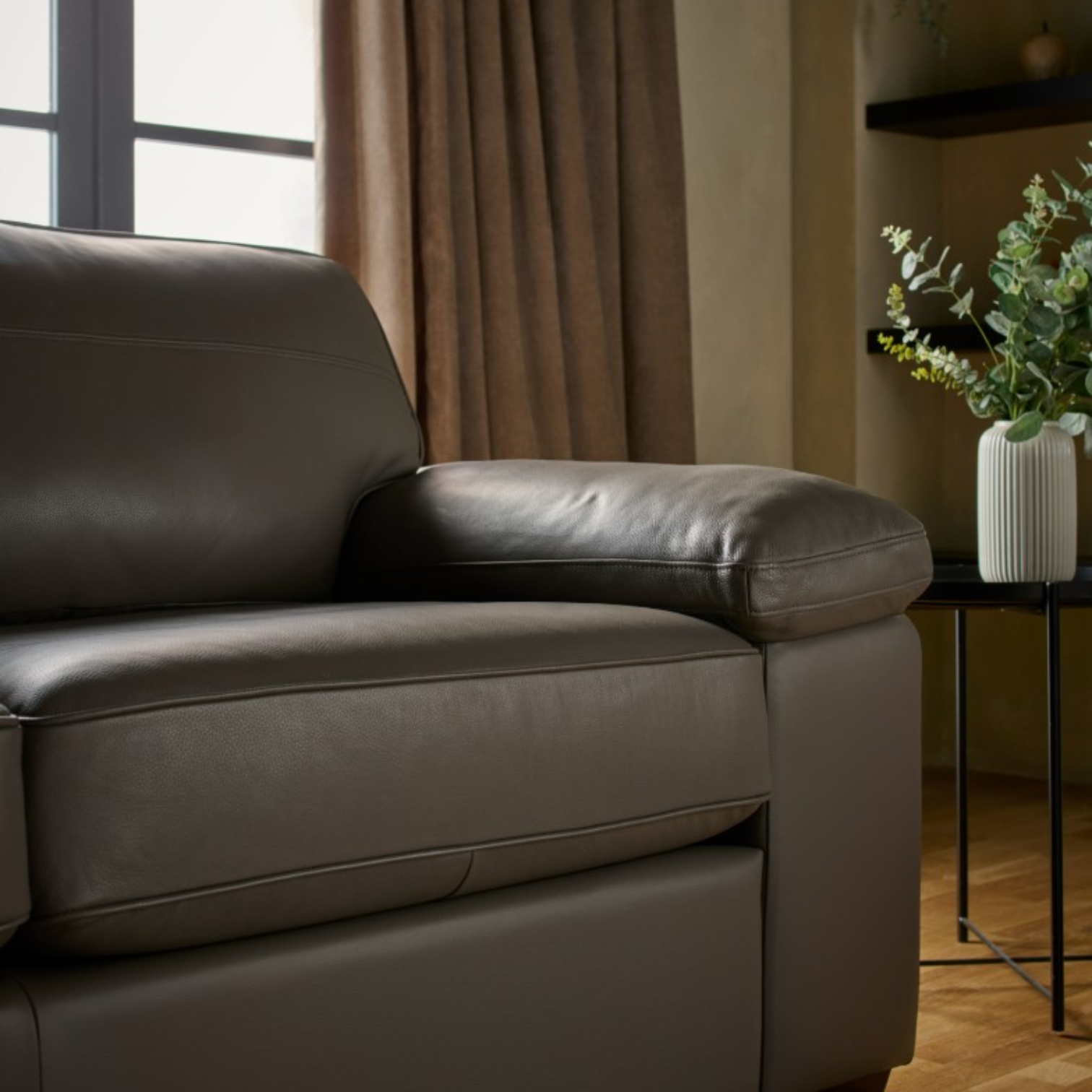 A close-up of a leather armchair in a styled living room setting with a side table, potted plant, and curtains.