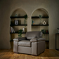 A modern leather  armchair is positioned in front of built-in dark wood shelves with plants and books, set against a textured beige wall with arched recesses.