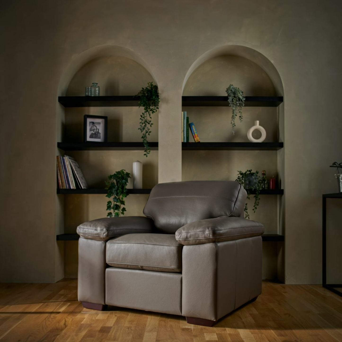 A modern leather  armchair is positioned in front of built-in dark wood shelves with plants and books, set against a textured beige wall with arched recesses.