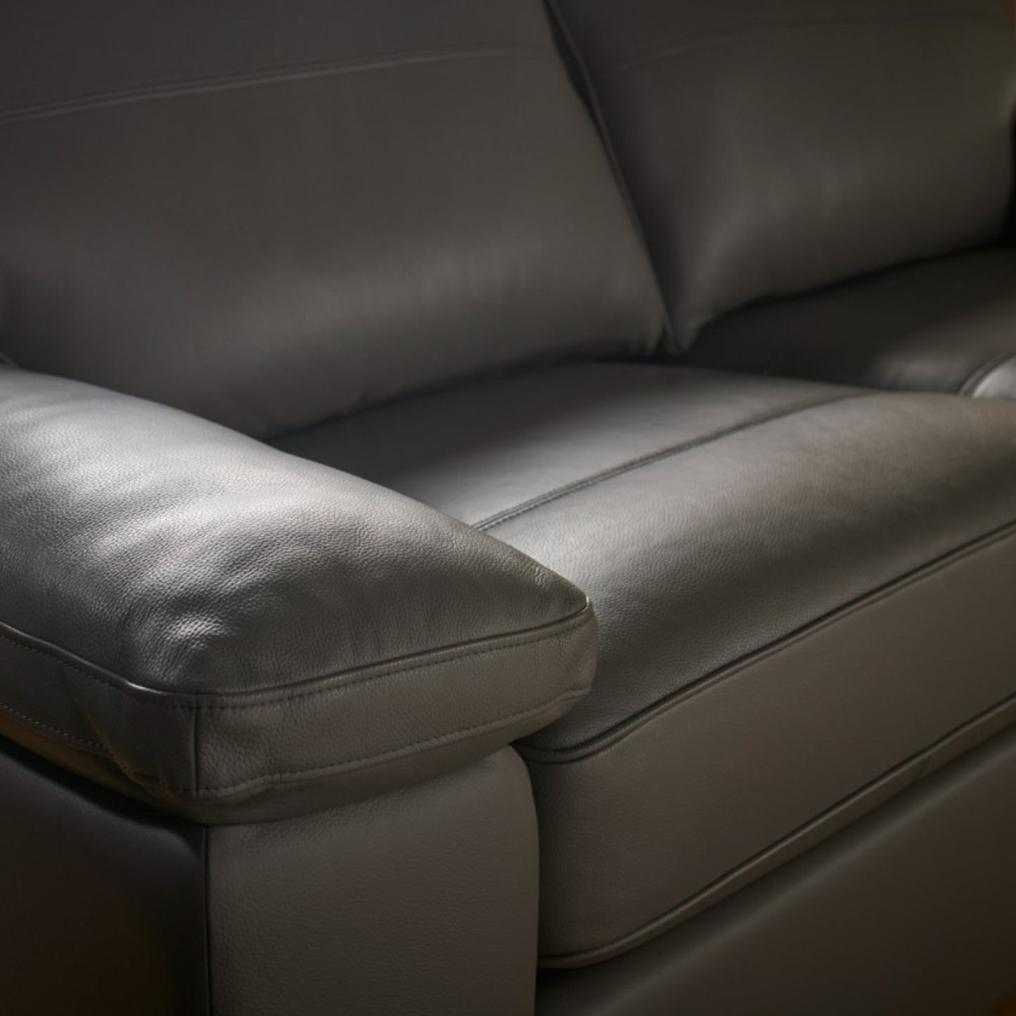 Close-up of a  leather sofa with padded armrest and seat cushions, showing the texture and stitching detail.