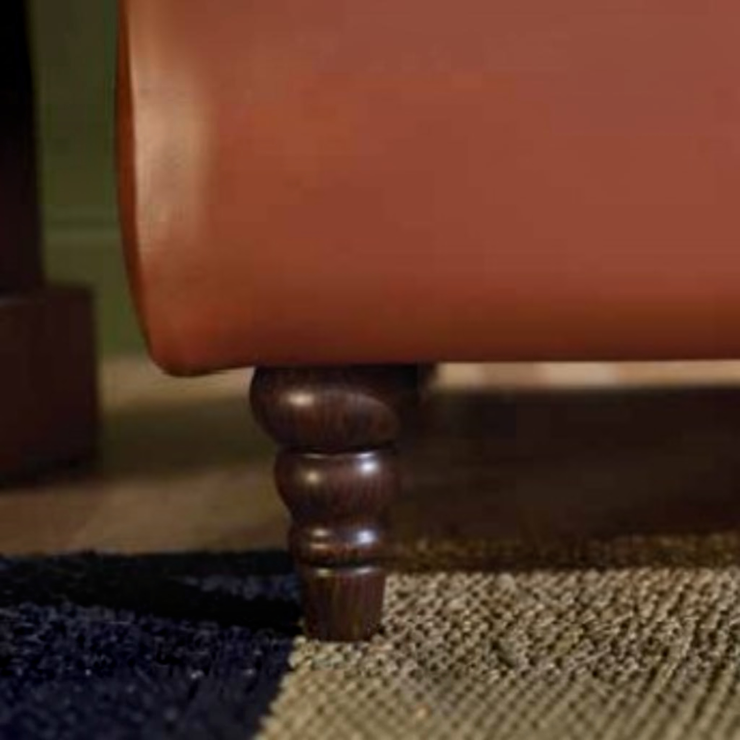 Close-up of a reddish-brown upholstered furniture piece with a turned dark wood leg, resting on a textured rug.