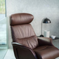 A brown leather recliner chair with a high headrest and padded backrest, shown in a modern interior setting with a side table, black lamp, and textured gray wall.