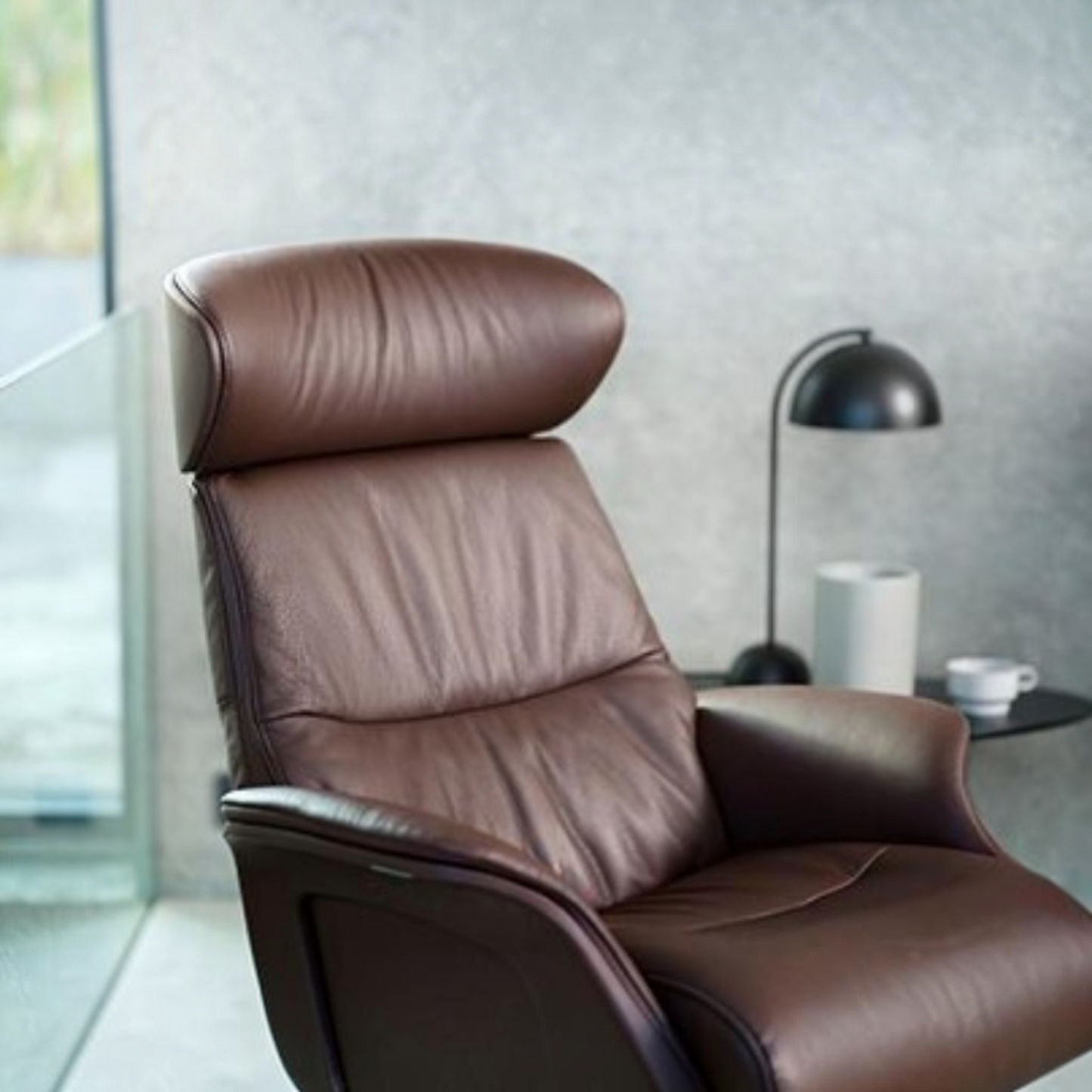 A brown leather recliner chair with a high headrest and padded backrest, shown in a modern interior setting with a side table, black lamp, and textured gray wall.