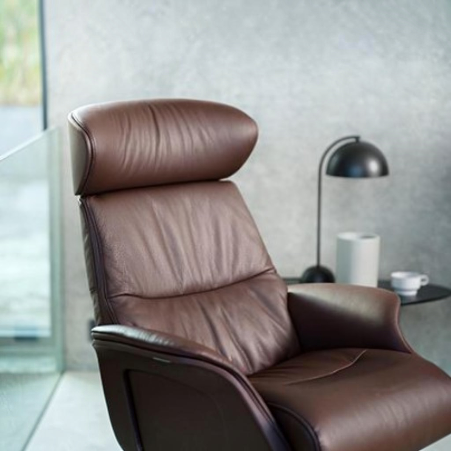 A brown leather recliner chair with a high headrest and padded backrest, shown in a modern interior setting with a side table, black lamp, and textured gray wall.