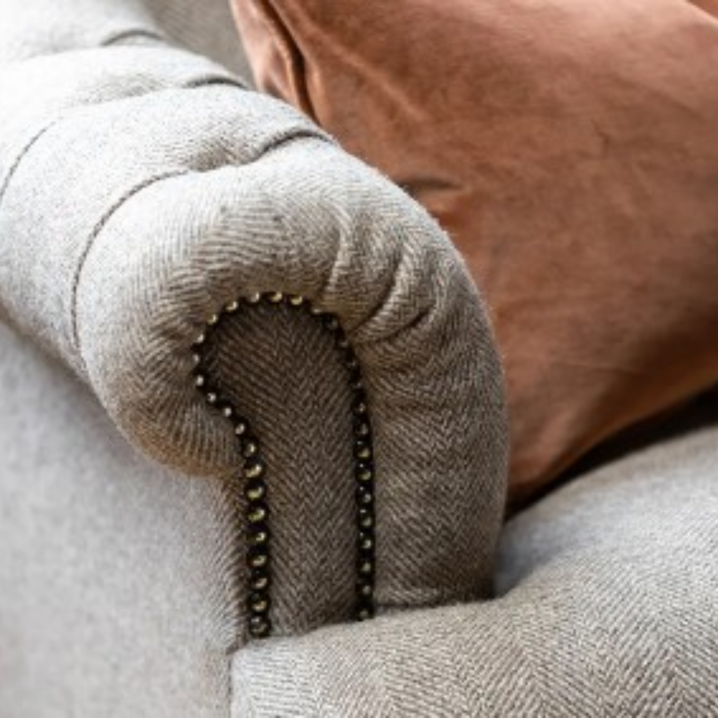 Close-up of a light gray herringbone upholstered armrest with dark nailhead trim, next to a brown velvet pillow.