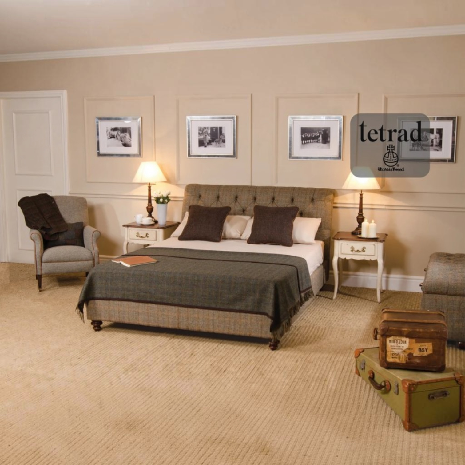 A staged bedroom interior featuring a tufted bed frame with a plaid bedding set, flanked by matching nightstands and lamps. A Harris Tweed upholstered armchair sits on the left, and vintage-style luggage rests on the floor on the right.