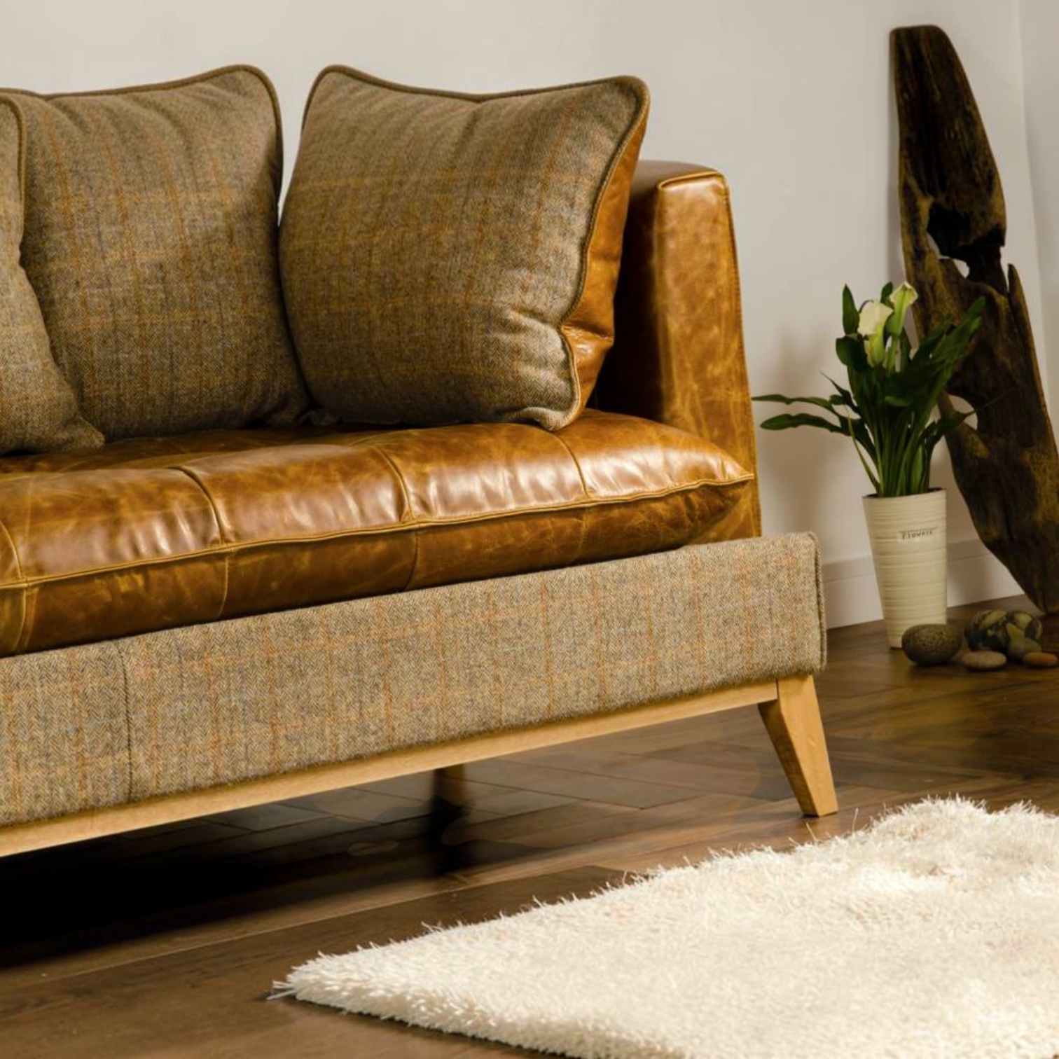 A close-up of a mid-century modern sofa featuring a brown leather seat and back cushions with a textured brown and beige tweed fabric base and backrest, styled in a living room setting with a white shag rug, a potted plant, and a wooden driftwood piece.