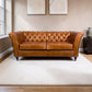 A classic Chesterfield sofa upholstered in warm Cerato brown leather, set on a large off-white rug in a living room with wood floors and neutral walls.