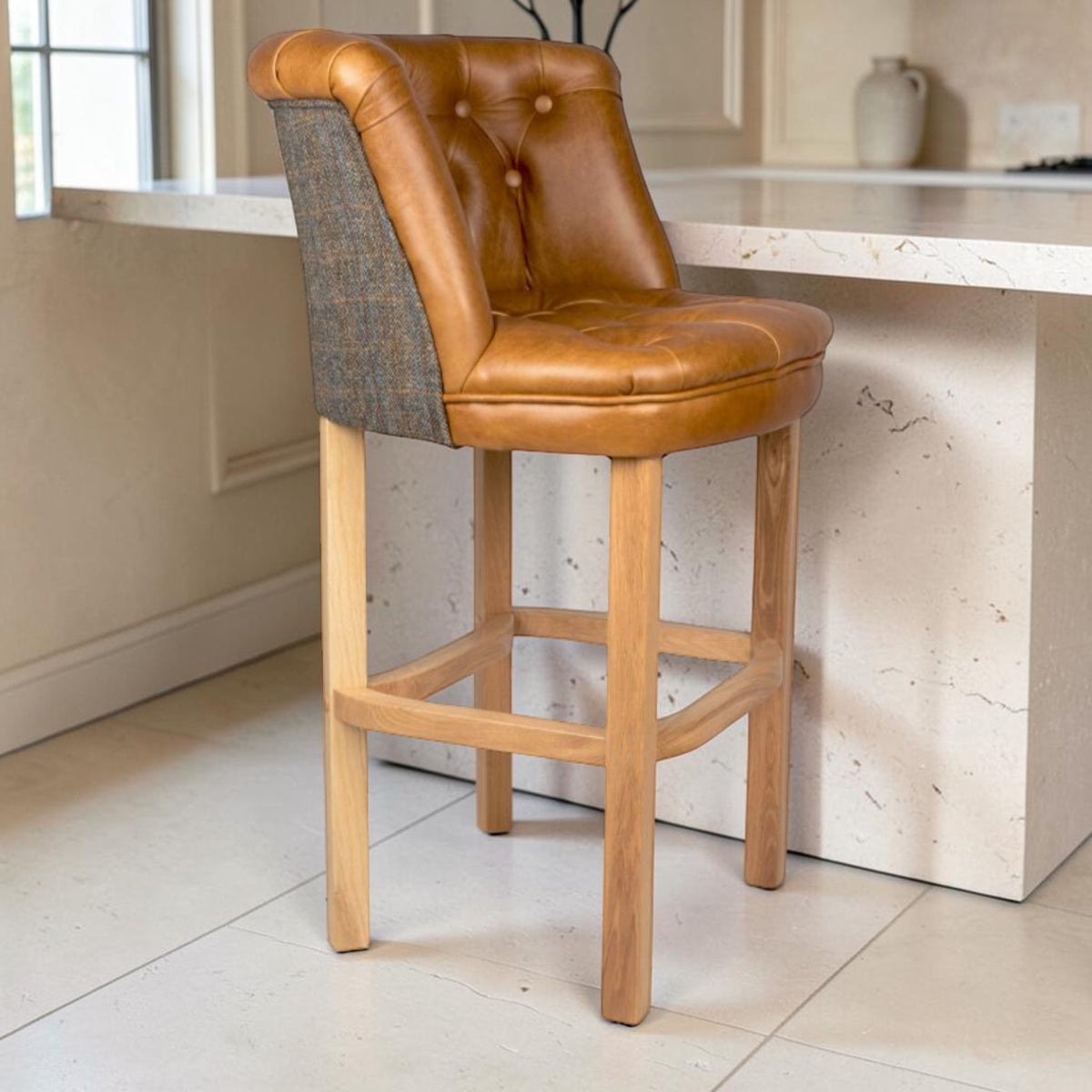 A wooden bar stool with a tufted Carato brown leather seat and backrest, featuring a Uist Harris Tweed side panel, set in a bright kitchen next to a marble island.
