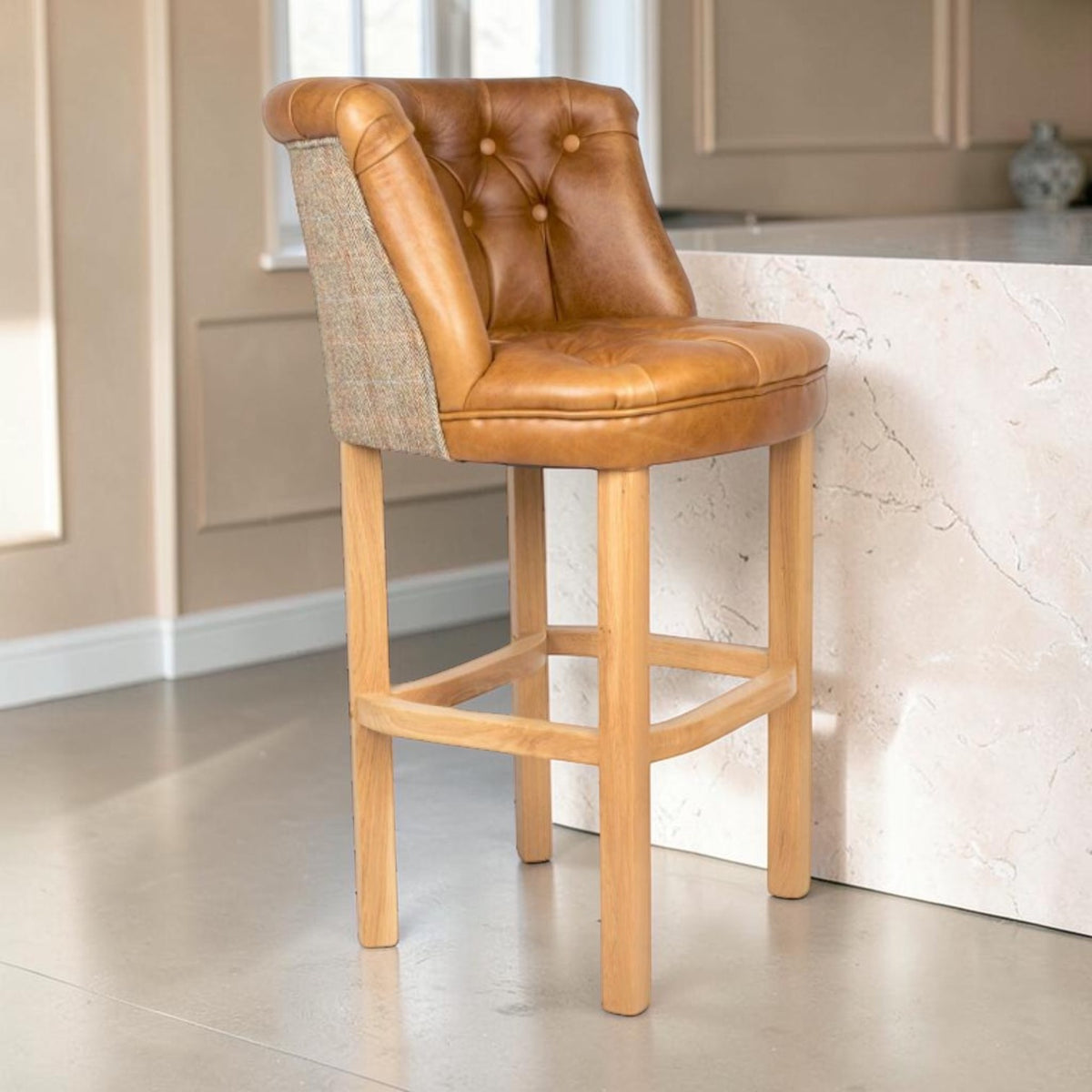 A counter-height bar stool with a tufted Cerato brown leather backrest and seat, combined with a textured beige Harris Tweed side panel and a light natural wood frame and legs, shown in a kitchen setting.