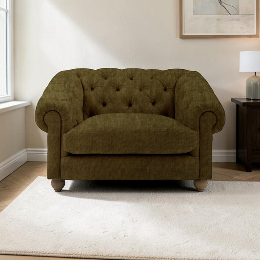 An olive green tufted armchair with rolled arms and wooden legs, positioned in a living room setting on a white rug next to a dark wood side table with a lamp.