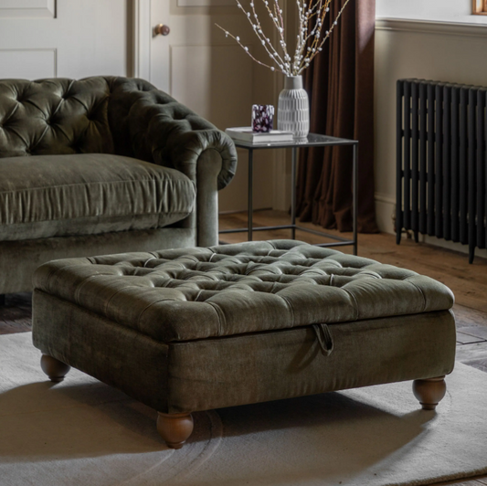 A large tufted storage ottoman in a muted olive green fabric, styled in a cozy living room next to a matching green sofa.