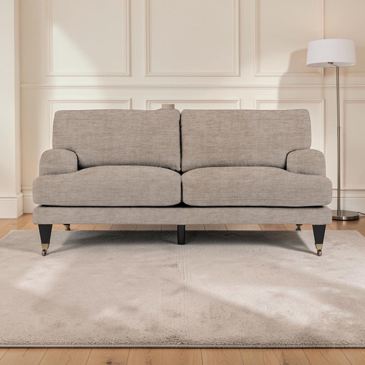 A upholstered sofa in a light beige textured fabric, positioned on a matching beige area rug in a bright living room with cream paneled walls, light wood floors, and a floor lamp.