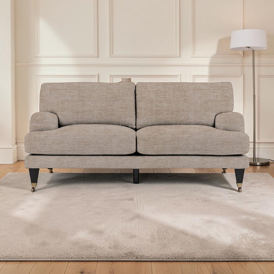 A upholstered sofa in a light beige textured fabric, positioned on a matching beige area rug in a bright living room with cream paneled walls, light wood floors, and a floor lamp.