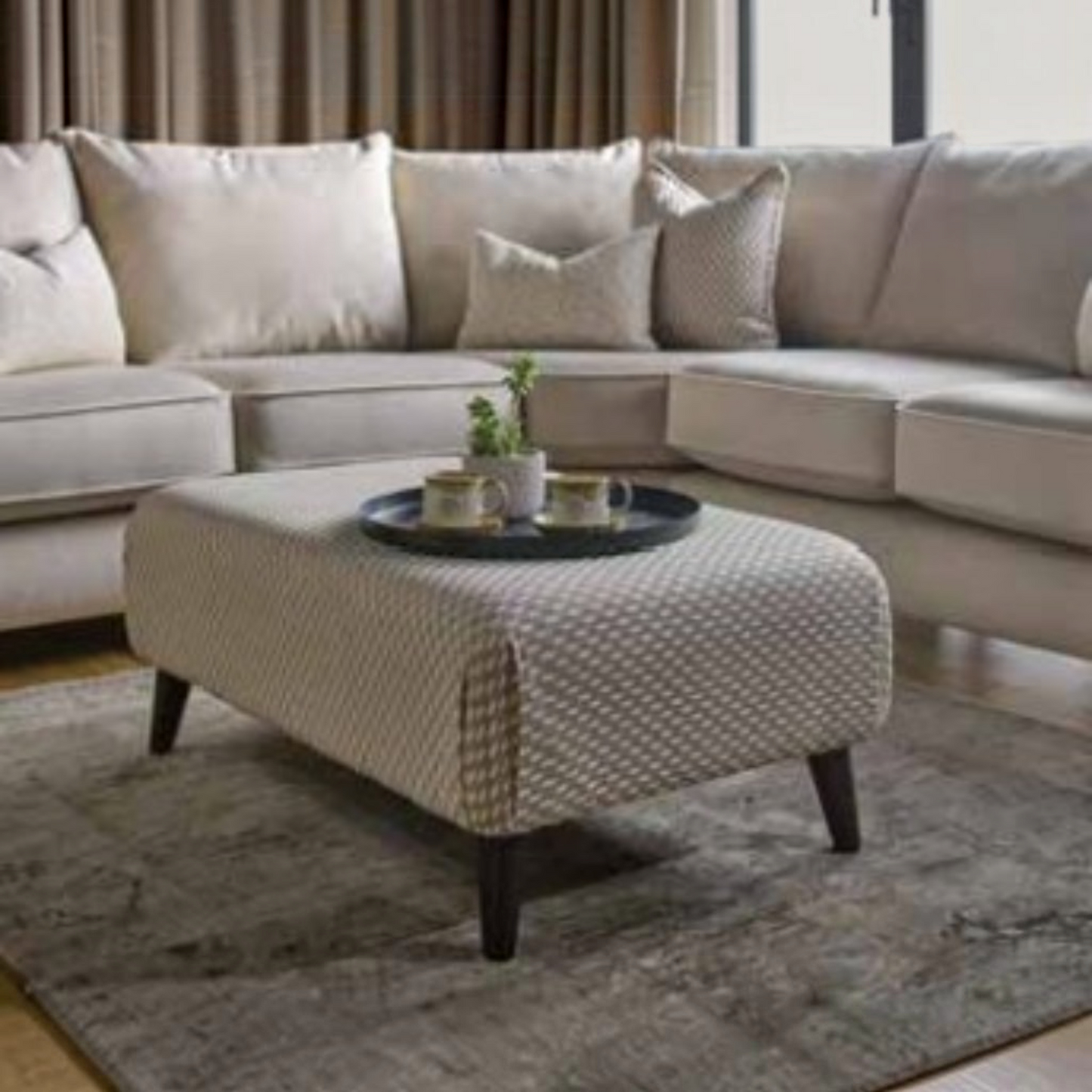 A modern living room scene featuring a light beige sectional sofa with matching throw pillows, a textured beige ottoman coffee table with dark wooden legs, and a gray area rug.