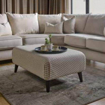 A modern living room scene featuring a light beige sectional sofa with matching throw pillows, a textured beige ottoman coffee table with dark wooden legs, and a gray area rug.