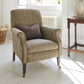 A classic English-style armchair upholstered in a textured tweed fabric with a brown accent pillow, set in a bright, sunlit room with white floorboards, a wooden side table, and a wicker basket.