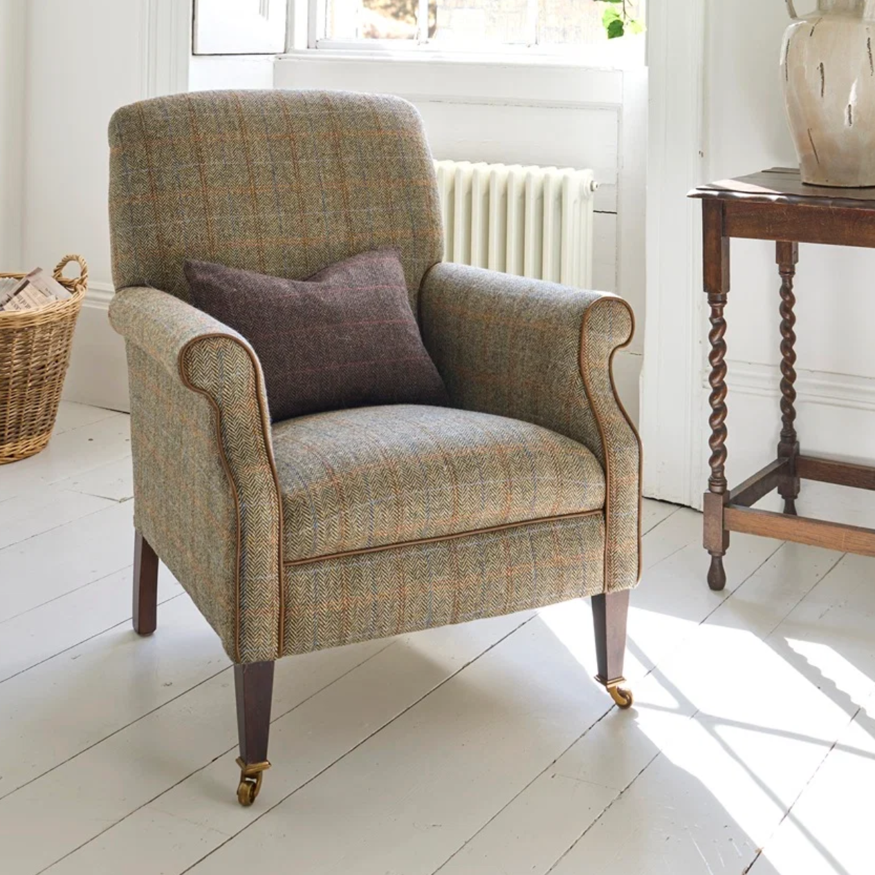 A classic English-style armchair upholstered in a textured tweed fabric with a brown accent pillow, set in a bright, sunlit room with white floorboards, a wooden side table, and a wicker basket.
