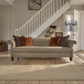A classic three-seater sofa in a warm beige and brown plaid tweed fabric, styled with dark brown and rust-orange throw pillows in a bright, modern living room with a staircase in the background. A Tetrad logo watermark is visible on the right.