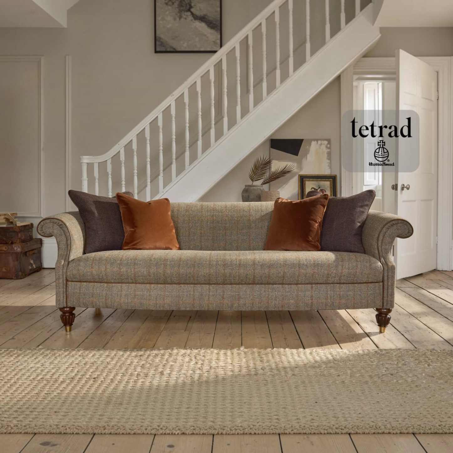 A classic three-seater sofa in a warm beige and brown plaid tweed fabric, styled with dark brown and rust-orange throw pillows in a bright, modern living room with a staircase in the background. A Tetrad logo watermark is visible on the right.