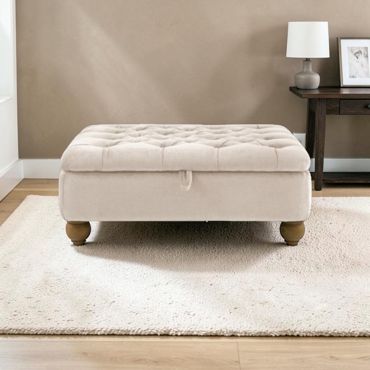 A cream tufted storage ottoman with wooden legs sits on a white shag rug in a living room setting with a beige wall and a dark wood side table.