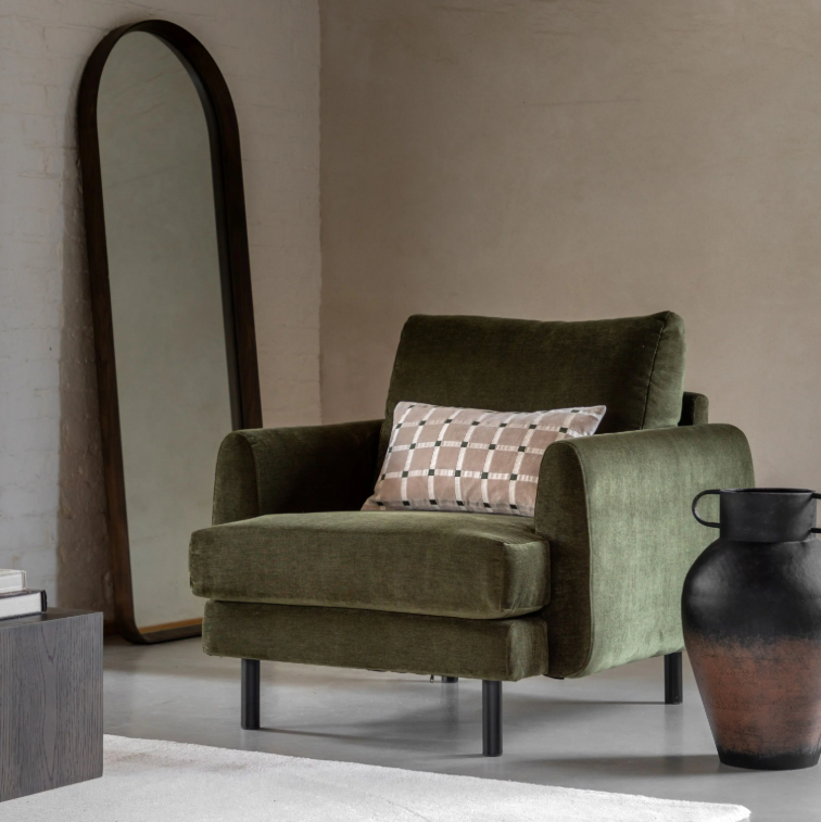 An olive green velvet armchair with a beige and brown checkered accent pillow, set in a minimalist room next to a large arched mirror and a rustic black-and-brown ceramic vase.