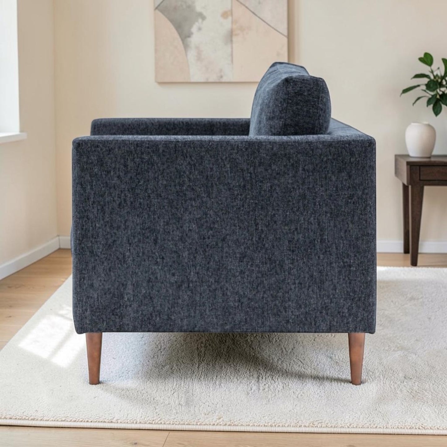 A side view of the Henley modern armchair upholstered in textured Charcoal fabric fabric with tapered wooden legs, shown in a styled living room setting on a white rug next to a side table.