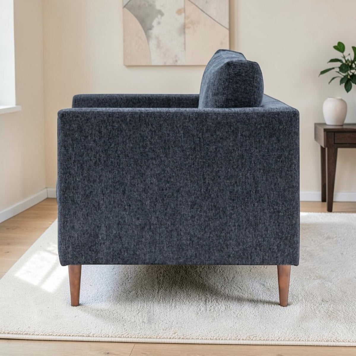 A side view of the Henley modern armchair upholstered in textured Charcoal fabric fabric with tapered wooden legs, shown in a styled living room setting on a white rug next to a side table.
