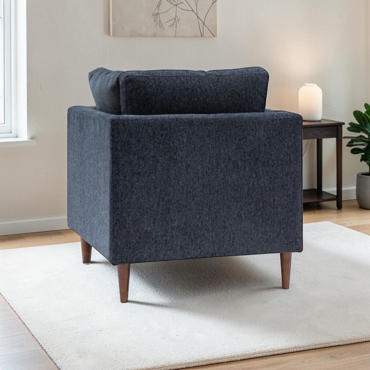 A back view of the Henley modern armchair upholstered in textured Charcoal fabric fabric with tapered wooden legs, shown in a styled living room setting on a white rug next to a side table.