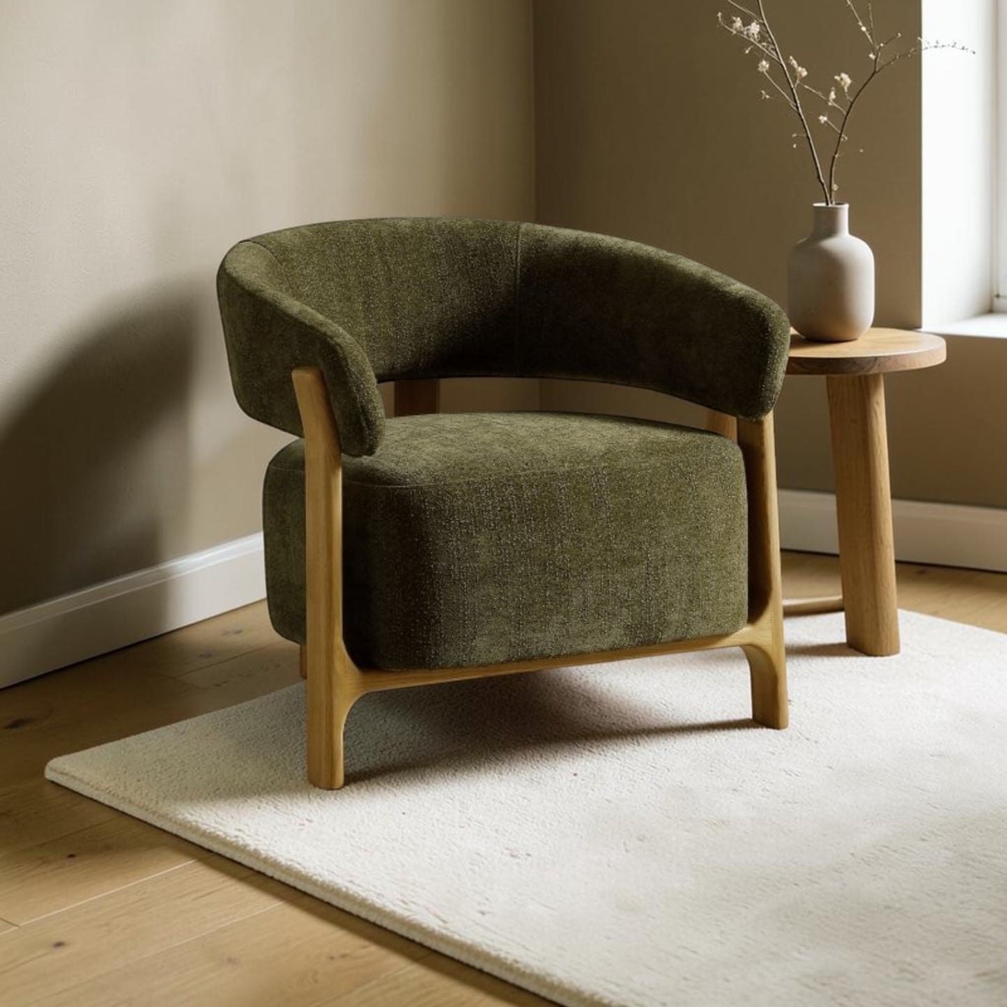 An olive green upholstered armchair with a curved back and light natural wood frame, styled in a minimalist room next to a small wooden side table with a vase.