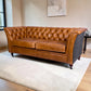 A classic Chesterfield sofa in warm tan leather with aUist Harris Tweed side panel, positioned on a white shag rug in a modern living room setting.